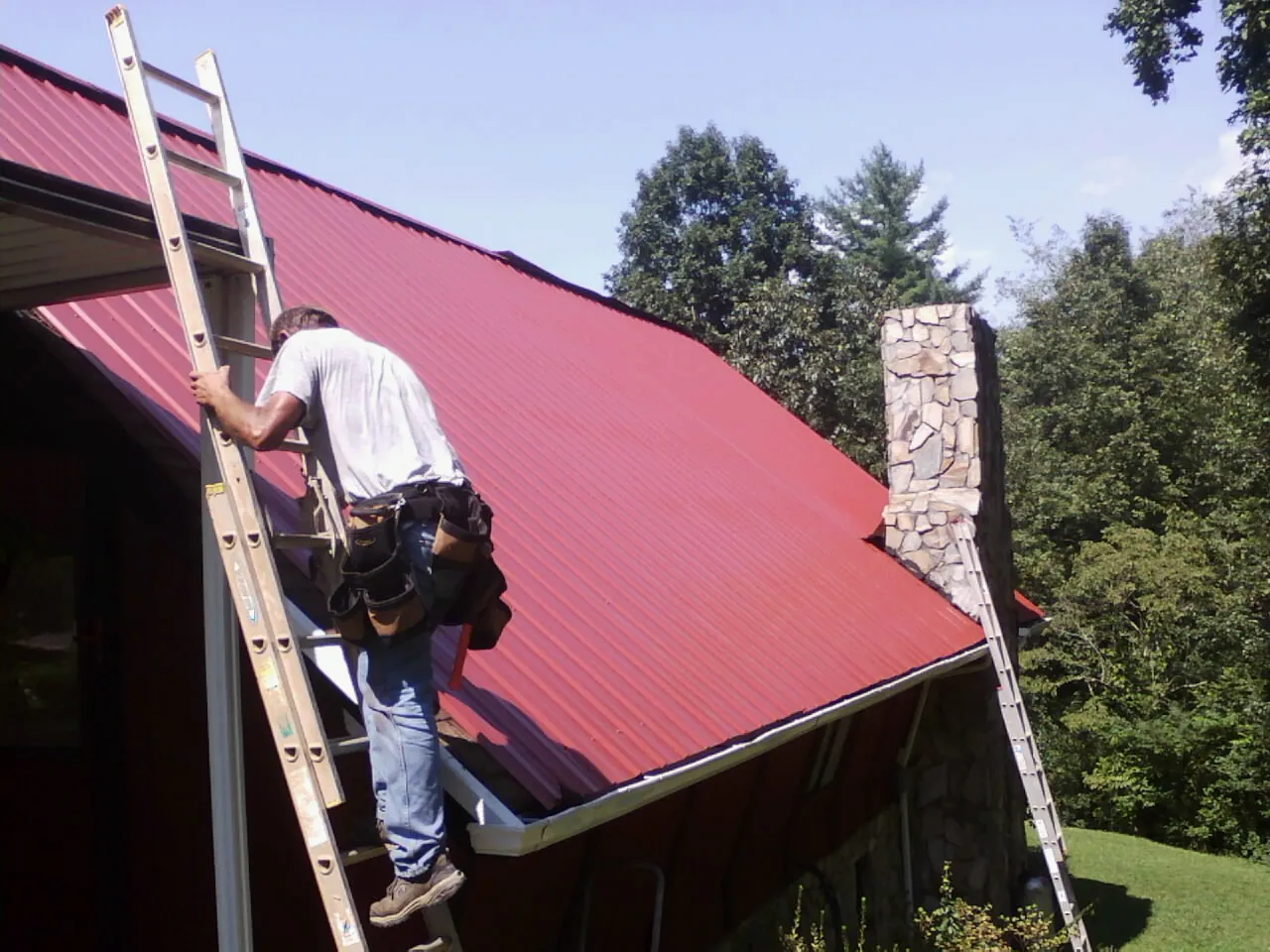 Roofer on a ladder beside a red metal roof and stone chimney in Andover